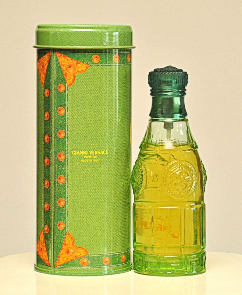 May include: A green perfume bottle and matching cylindrical container. The bottle is clear with a green cap and base, filled with yellow liquid. The container has a denim-like texture with orange and gold accents. The text "GIANNI VERSACE PROFUMI MADE IN ITALY" is visible.