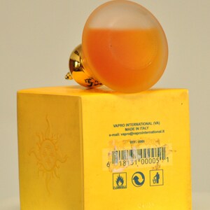 May include: Yellow box with a sun graphic and text that reads "VAPRO INTERNATIONAL (VA) MADE IN ITALY e-mail: vapro@vaprointernational.it REF: 0005 6 18131 00005 1 PLAMMABLE". A clear glass bottle with a gold cap and orange liquid is on top of the box.