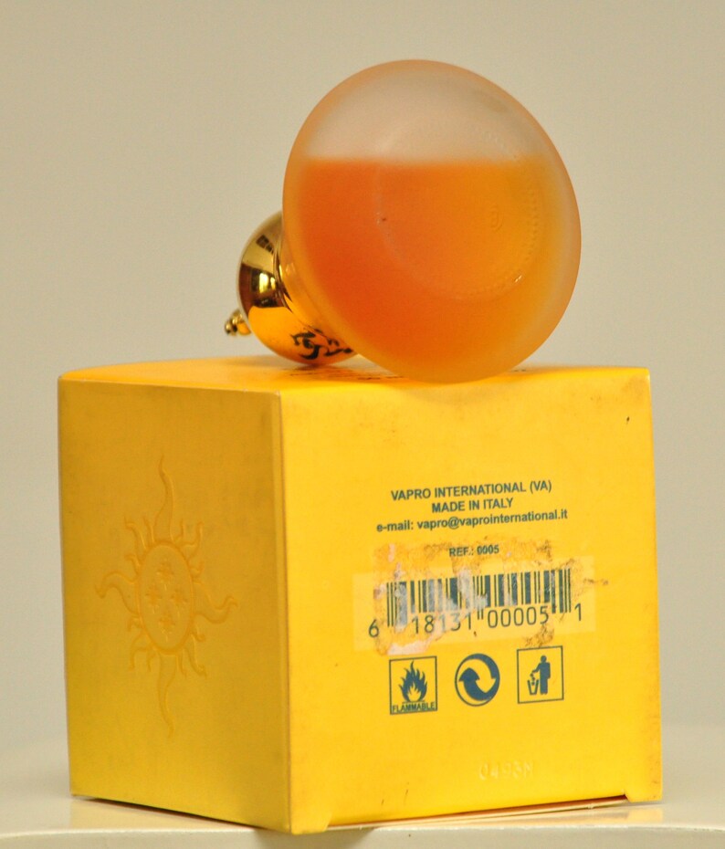 May include: Yellow box with a sun graphic and text that reads "VAPRO INTERNATIONAL (VA) MADE IN ITALY e-mail: vapro@vaprointernational.it REF: 0005 6 18131 00005 1 PLAMMABLE". A clear glass bottle with a gold cap and orange liquid is on top of the box.