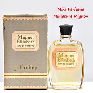 Può includere: Un set vintage di bottiglia e scatola di profumo. La bottiglia è trasparente con un tappo nero e contiene un liquido giallo chiaro. La scatola è grigia con un'etichetta rosa e oro che recita "Muguet Elisabeth Eau de Toilette" e "J. Collins". È visibile anche il testo "Mini Perfume Miniature Mignon".