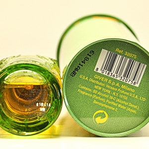May include: A green glass perfume bottle with a textured design, next to a green cylindrical container. The bottle contains a golden-colored liquid. The container has a barcode and text including "Versace Profumi".