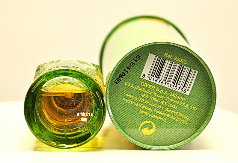 May include: A green glass perfume bottle with a textured design, next to a green cylindrical container. The bottle contains a golden-colored liquid. The container has a barcode and text including "Versace Profumi".