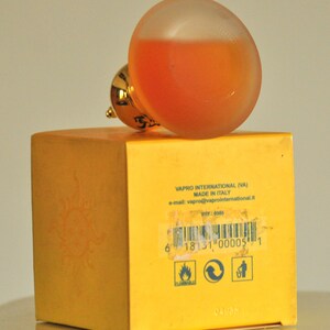 May include: A yellow box with a sun graphic and the text "VAPRO INTERNATIONAL (VA) MADE IN ITALY e-mail: vapro@vaprointernational.it REF: 0005 6 18131 000051 0498M" printed on it. A clear glass bottle with a gold cap and orange liquid inside is sitting on top of the box.