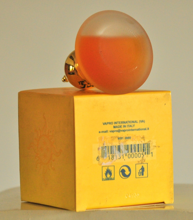 May include: A yellow box with a sun graphic and the text "VAPRO INTERNATIONAL (VA) MADE IN ITALY e-mail: vapro@vaprointernational.it REF: 0005 6 18131 000051 0498M" printed on it. A clear glass bottle with a gold cap and orange liquid inside is sitting on top of the box.