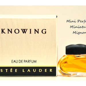 May include: A miniature perfume bottle and box set. The box is beige with the word "KNOWING" in black. The perfume bottle is a small, rounded shape with a black cap, filled with amber-coloured liquid. The text "ESTEE LAUDER" is on the box.