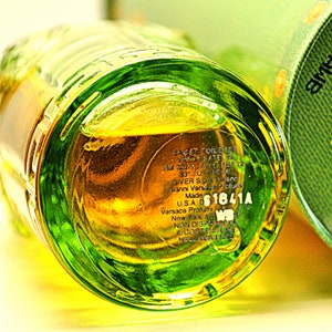 May include: Close-up of a clear glass perfume bottle with a green rim and a yellow liquid inside. The bottom of the bottle has printed text, including the brand name and a product code. The bottle is partially visible.