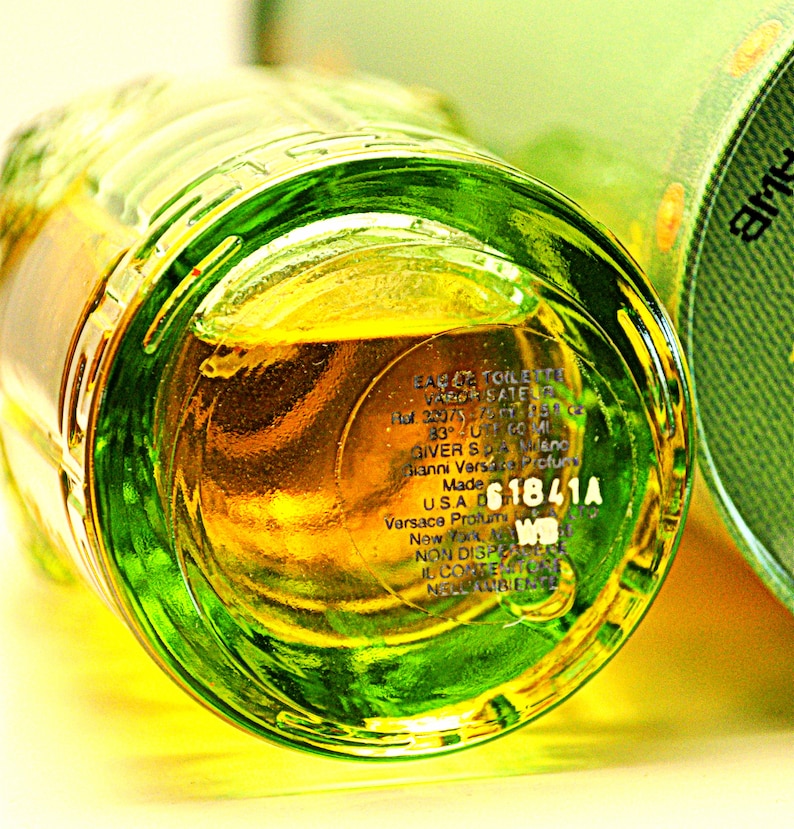 May include: Close-up of a clear glass perfume bottle with a green rim and a yellow liquid inside. The bottom of the bottle has printed text, including the brand name and a product code. The bottle is partially visible.