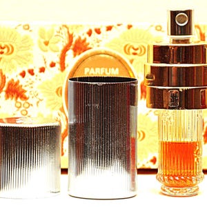 Può includere: Un flacone di profumo in argento e vetro con un design a coste. Il flacone è etichettato "PARFUM" e si trova su una scatola con un motivo.