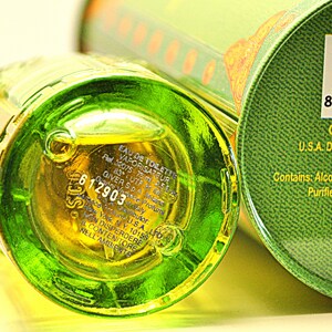 May include: Close-up of a green glass perfume bottle with a yellow liquid inside. The bottle is partially visible, with text and a product label. A green circular container is also in view, with a barcode and text.