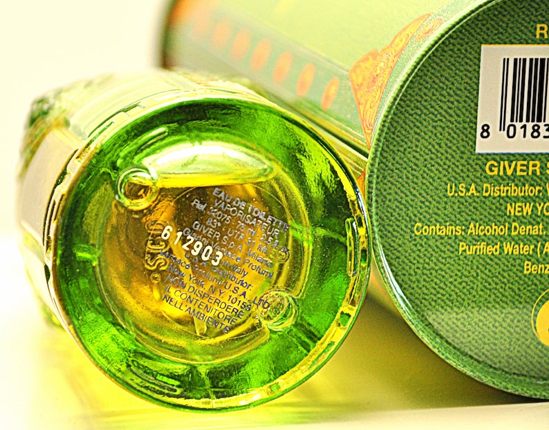 May include: Close-up of a green glass perfume bottle with a yellow liquid inside. The bottle is partially visible, with text and a product label. A green circular container is also in view, with a barcode and text.