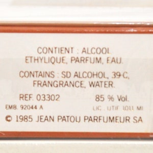 May include: A white box with brown trim and text in French and English. The text reads "CONTIENT: ALCOOL ETHYLIQUE, PARFUM, EAU. CONTAINS: SD ALCOHOL, 39-C, FRANGRANCE, WATER. REF. 03302 EMB 92044 A 85% Vol. LIC. UTIF 1011 MI 1985 JEAN PATOU PARFUMEUR SA".