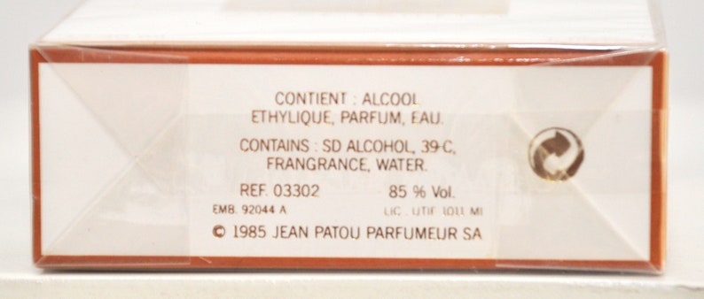 May include: A white box with brown trim and text in French and English. The text reads "CONTIENT: ALCOOL ETHYLIQUE, PARFUM, EAU. CONTAINS: SD ALCOHOL, 39-C, FRANGRANCE, WATER. REF. 03302 EMB 92044 A 85% Vol. LIC. UTIF 1011 MI 1985 JEAN PATOU PARFUMEUR SA".