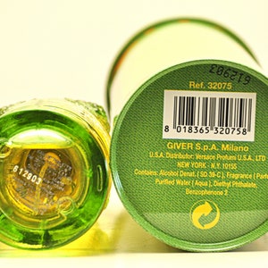 May include: A green glass perfume bottle with a textured design, next to a green and white cylindrical box. The bottle contains a yellow liquid. The box has a barcode and text that includes "GIVER S.p.A. Milano".