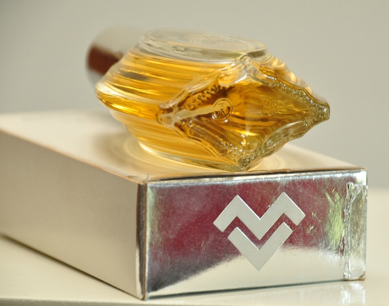 K&ouml;nnte beinhalten: Eine durchsichtige Glasflasche mit Parfum, einem goldenen Verschluss und einer silbernen Schachtel mit einem wellenf&ouml;rmigen Design. Auf der Schachtel steht "Aquarius" gedruckt.