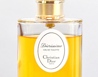 Könnte beinhalten: Eine rechteckige Glasflasche Diorissimo Eau de Toilette. Die Flasche hat einen klaren, abgerundeten Verschluss und einen goldfarbenen Hals. Die Flüssigkeit im Inneren ist goldgelb, und ein weißes ovales Etikett zeigt den Markennamen und Details.