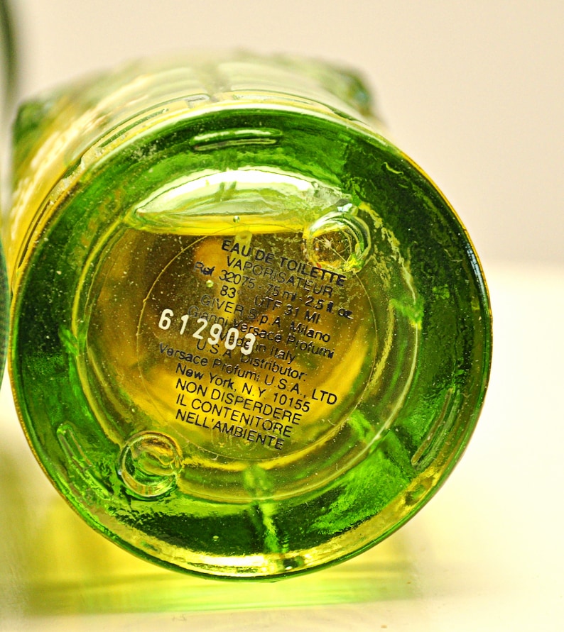 May include: Close-up of a green glass perfume bottle base with yellow liquid inside. The bottom of the bottle has printed text including "EAU DE TOILETTE" and "75 ml - 2.5 fl oz". The bottle is labeled "Versace Profumi".