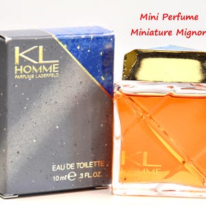 Peut inclure: Un flacon miniature de parfum KL Homme Eau de Toilette, 10 ml, à côté de sa boîte. La boîte est gris foncé avec un accent bleu et or. Le flacon de parfum est orange et a un bouchon doré. Le texte "KL HOMME" est visible.