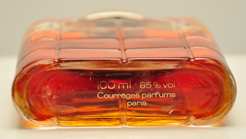 Puede incluir: Una botella de perfume de vidrio transparente con una tapa dorada. La botella est&aacute; llena de un l&iacute;quido marr&oacute;n rojizo. La etiqueta de la botella dice "100 ml 85% vol Courr&egrave;ges parfums paris".