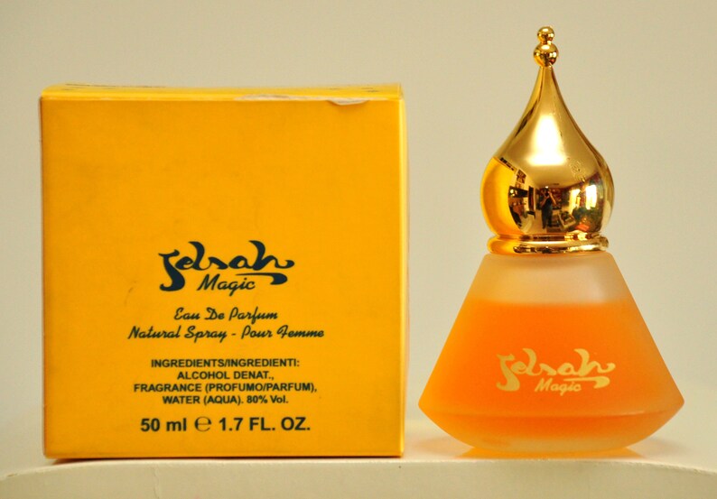May include: A yellow box with black text that reads "Sarah Magic Eau De Parfum Natural Spray - Pour Femme" and "INGREDIENTS/INGREDIENTI: ALCOHOL DENAT., FRAGRANCE (PROFUMO/PARFUM), WATER (AQUA). 80% Vol. 50 ml e 1.7 FL. OZ."  Next to the box is a clear glass bottle with a gold top and a yellow liquid inside. The bottle has black text that reads "Sarah Magic".