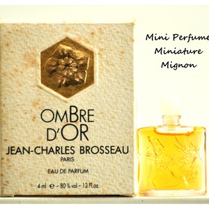 Può includere: Un set di bottiglia e scatola di profumo in miniatura. La scatola è beige con dettagli dorati e le parole "OMBRE D'OR" e "JEAN-CHARLES BROSSEAU PARIS" stampate sopra. Il flacone di profumo è quadrato, riempito di liquido giallo e ha un tappo trasparente. È visibile anche il testo "Mini Perfume Miniature Mignon".