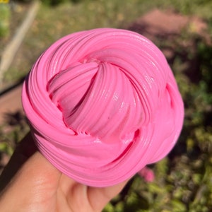 May include: A close-up of a pink slime, a popular sensory toy, with a smooth, glossy finish. The slime is being held in a hand, and the background is blurred.