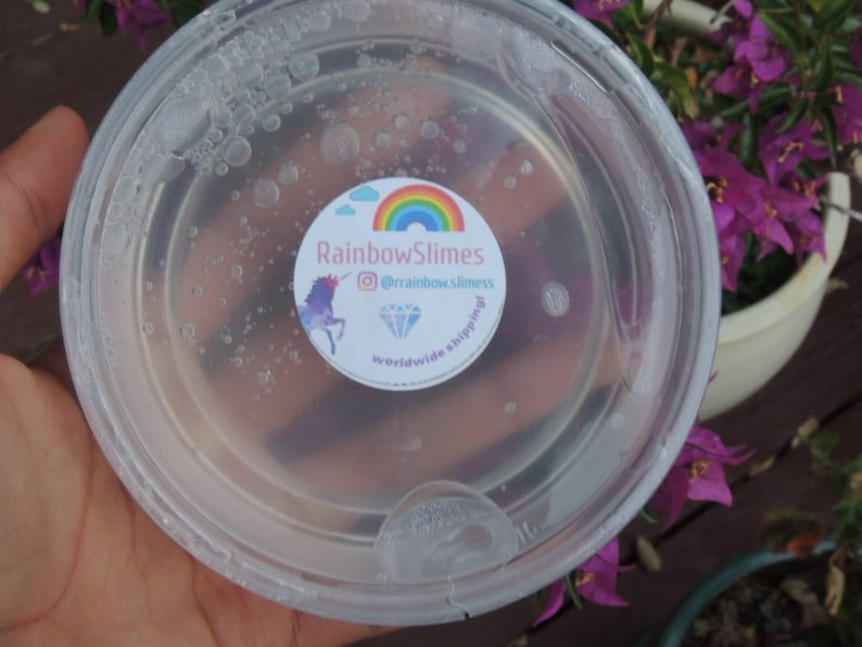 Puede incluir: Un recipiente de pl&aacute;stico transparente lleno de slime transparente con una pegatina en el lateral que dice "Rainbow Slimes @rainbow.slimes worldwide shipping".