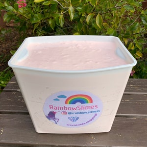Pode incluir: Um grande recipiente branco cheio de slime rosa. O recipiente tem um adesivo no lado que diz "Rainbow Slimes" com um gráfico de unicórnio e arco-íris. O adesivo também inclui o texto "@rainbow.slimes" e "envio mundial."