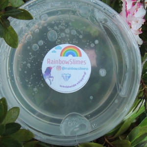 Puede incluir: Una slime transparente en un recipiente de pl&aacute;stico con una pegatina que dice "Rainbow Slimes" con un arco&iacute;ris, un unicornio, un diamante y el texto "@rainbow.slimes worldwide shipping!"