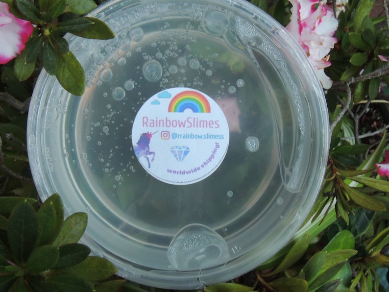 Puede incluir: Una slime transparente en un recipiente de pl&aacute;stico con una pegatina que dice "Rainbow Slimes" con un arco&iacute;ris, un unicornio, un diamante y el texto "@rainbow.slimes worldwide shipping!"