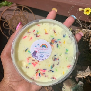 May include: A clear plastic container filled with a light yellow slime with colorful sprinkles and a white sticker that says "RainbowSlimes @rainbowslimes made in the USA".