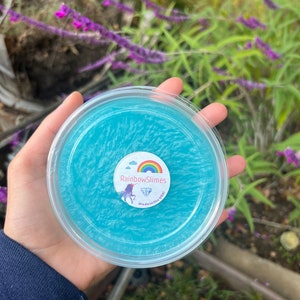 Könnte beinhalten: Ein klarer Plastikbehälter gefüllt mit hellblauem Schleim. Der Behälter hat ein rundes Etikett mit einem Regenbogen, einem Einhorn und dem Text "RainbowSlimes Made in the USA". Der Schleim hat ein schimmerndes, strukturiertes Aussehen.