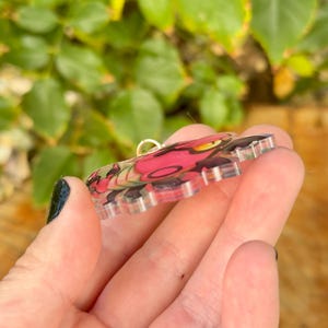 Puede incluir: Un colgante de acr&iacute;lico transparente con un dise&ntilde;o rosa y negro, con detalles en amarillo. El colgante tiene un lazo dorado para colgar. El dise&ntilde;o parece ser una imagen estilizada de una criatura. El colgante est&aacute; en la mano de una persona.