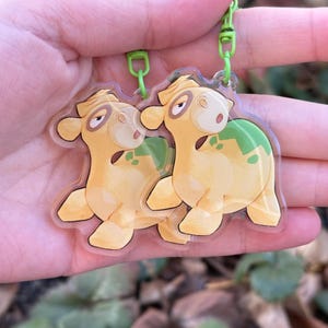 May include: Two clear acrylic keychains featuring a cartoon camel design. The camels are yellow with green accents and a green clasp. The keychains are held up against a blurred background of green leaves.