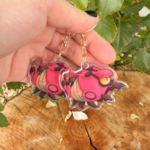 Puede incluir: Dos llaveros de acr&iacute;lico transparente con una criatura rosa de dibujos animados con ojos amarillos y detalles oscuros. Los llaveros tienen cadenas y cierres dorados. El dise&ntilde;o de la criatura incluye una forma circular y un cuerpo segmentado.