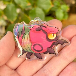 Puede incluir: Un llavero de acr&iacute;lico transparente con una criatura de dibujos animados roja y negra con un gran ojo amarillo. La criatura tiene un cuerpo segmentado, verde y rosa iridiscente. Un peque&ntilde;o anillo dorado est&aacute; unido en la parte superior.