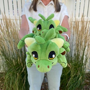 Puede incluir: Dos peluches de dragón verde con grandes ojos negros, detalles amarillos y garras marrones. Los dragones están apilados, uno encima del otro. Los juguetes son suaves y tiernos, perfectos para niños.