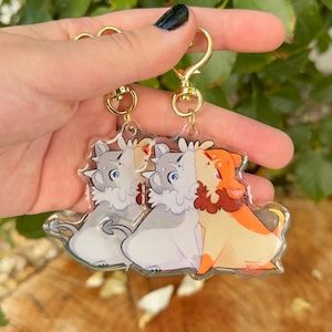 May include: Clear acrylic keychains featuring cartoon dogs in gray, white, and orange. Each keychain has a gold-colored clasp. The dogs are in a hugging pose, with one dog kissing another.