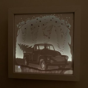 May include: A white framed shadow box with a silhouette of a pickup truck carrying a Christmas tree. The truck is driving through a snowy forest with stars in the sky.
