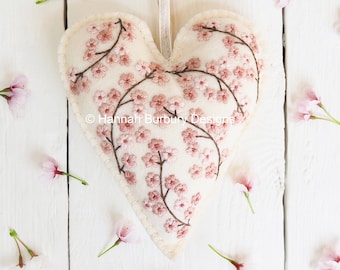 May include: A white felt heart-shaped ornament embroidered with pink cherry blossoms and branches. The ornament is surrounded by pink cherry blossoms on a white wooden background.