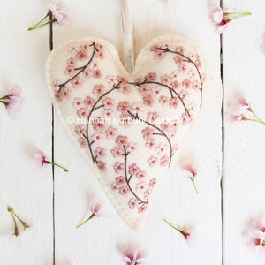 May include: A white felt heart-shaped ornament embroidered with pink cherry blossoms and branches. The ornament is surrounded by pink cherry blossoms on a white wooden background.