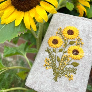 May include: A close-up of a light gray felt item with an embroidered sunflower design. The embroidery features yellow sunflowers with brown centers and green leaves. The background includes vibrant yellow sunflowers and green foliage.