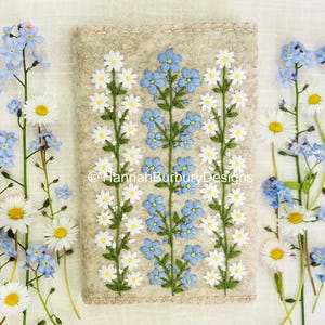 May include: A beige felt pouch with embroidered white and blue forget-me-not flowers. The pouch is surrounded by real forget-me-not flowers and daisies.