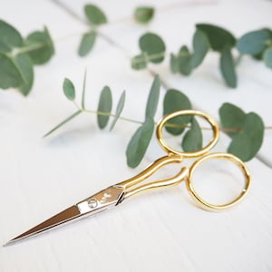 May include: Small gold-toned scissors with a silver blade. The scissors are on a white surface with green eucalyptus leaves.
