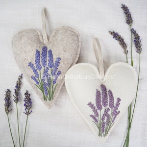 May include: Two felt heart-shaped lavender sachets with embroidered sprigs of lavender. The sachets have a loop for hanging and are a light beige colour.