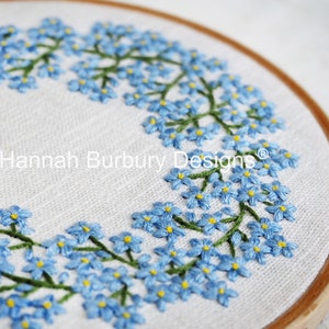 May include: A close-up of a circular embroidery hoop with a white linen fabric background. The fabric is embroidered with a wreath of blue forget-me-not flowers with green stems. The text "Hannah Burbury Designs" is embroidered in the center of the hoop.