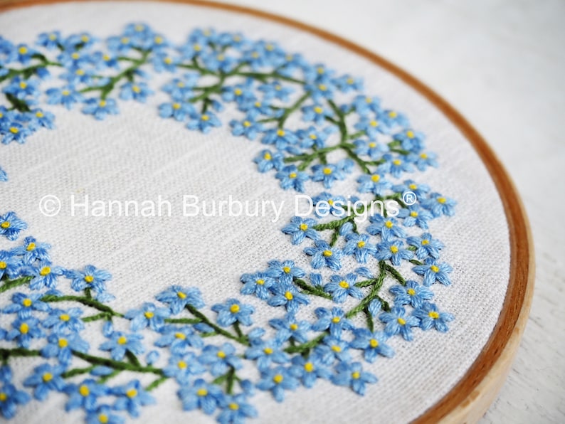 May include: A close-up of a circular embroidery hoop with a white linen fabric background. The fabric is embroidered with a wreath of blue forget-me-not flowers with green stems. The text "Hannah Burbury Designs" is embroidered in the center of the hoop.