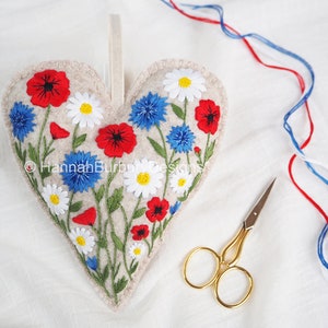 May include: A felt heart-shaped ornament with embroidered red poppies, blue cornflowers, and white daisies. The ornament has a loop for hanging and is surrounded by white fabric.