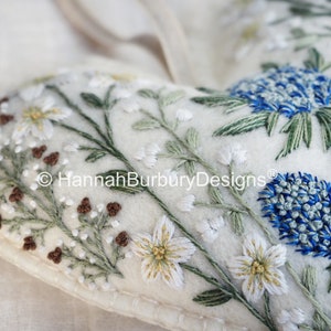 May include: A white felt heart-shaped cushion with embroidered flowers and leaves in shades of green, white, and brown. The cushion has a blue and white flower in the top right corner. The text "&copy; HannahBurburyDesigns&reg;" is embroidered in the centre of the cushion.