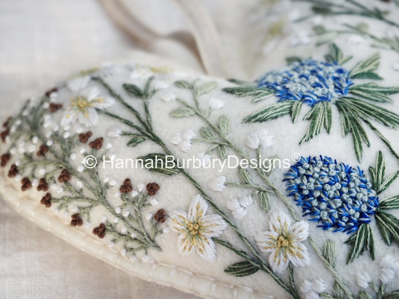 May include: A white felt heart-shaped cushion with embroidered flowers and leaves in shades of green, white, and brown. The cushion has a blue and white flower in the top right corner. The text "&copy; HannahBurburyDesigns&reg;" is embroidered in the centre of the cushion.