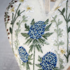 May include: White felt heart-shaped ornament with blue and green embroidered flowers. The ornament has a white ribbon loop for hanging. The text "Hannah Burbury Designs" is embroidered on the ornament.